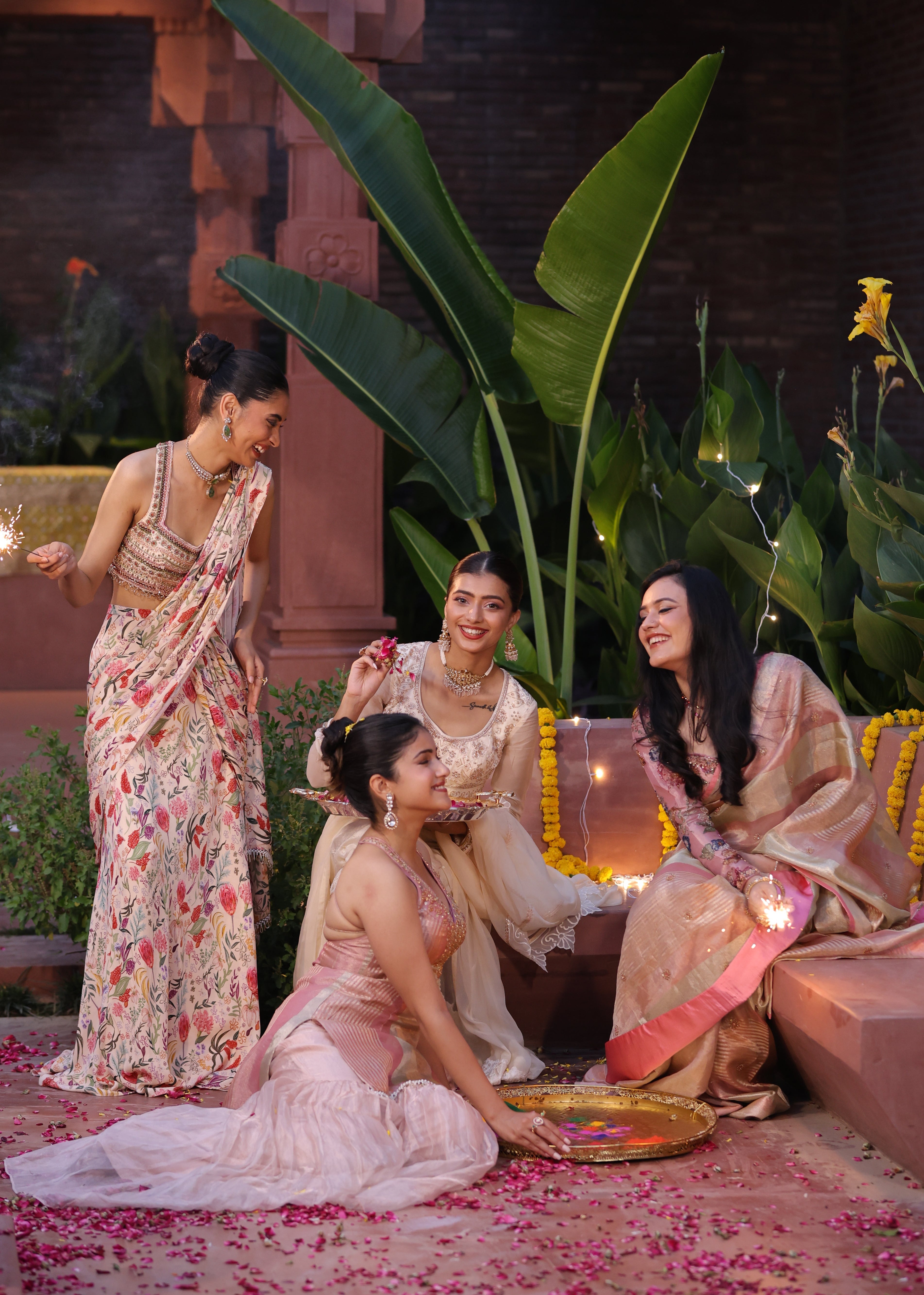 Three women in traditional attire sitting on a floral-patterned carpet with plants in the foreground.